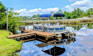 A pontoon boat docked at a wooden pier with a canopy, surrounded by lush greenery and calm water. - Boat Included Pool Pet Friendly Most Unique & Intimate Beachfront By Boat Home! (Pensacola)