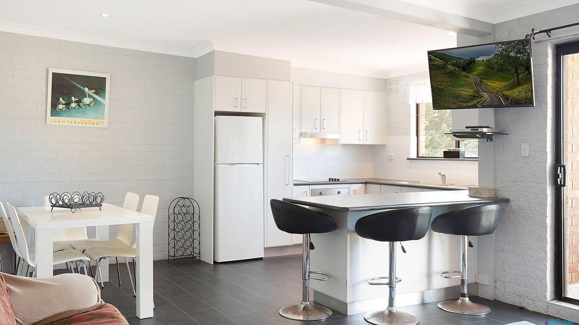 Une cuisine moderne avec des meubles blancs, un réfrigérateur, une table à manger avec des chaises et un coin bar avec des tabourets.
