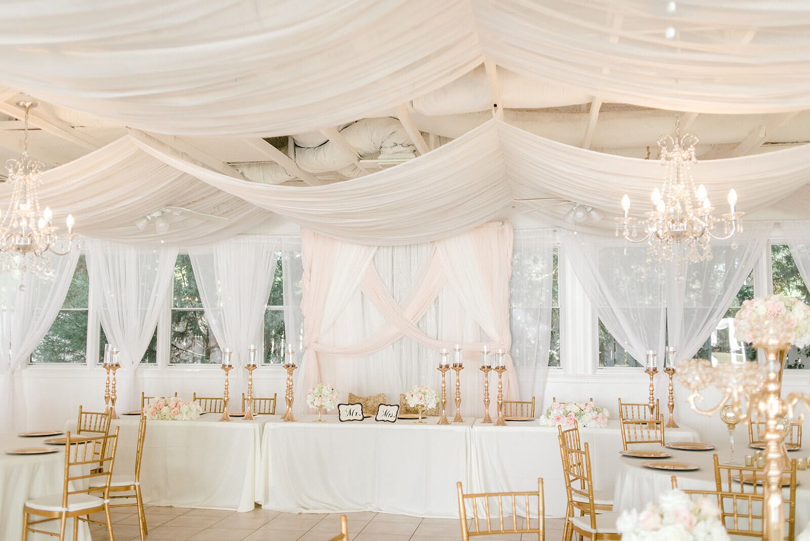A wedding reception area with white draped ceilings, chandeliers, and gold chairs.