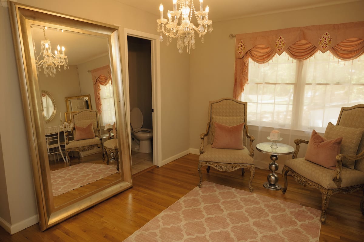 A room with a large mirror, a chandelier, two armchairs, a small round table, and a window with curtains.