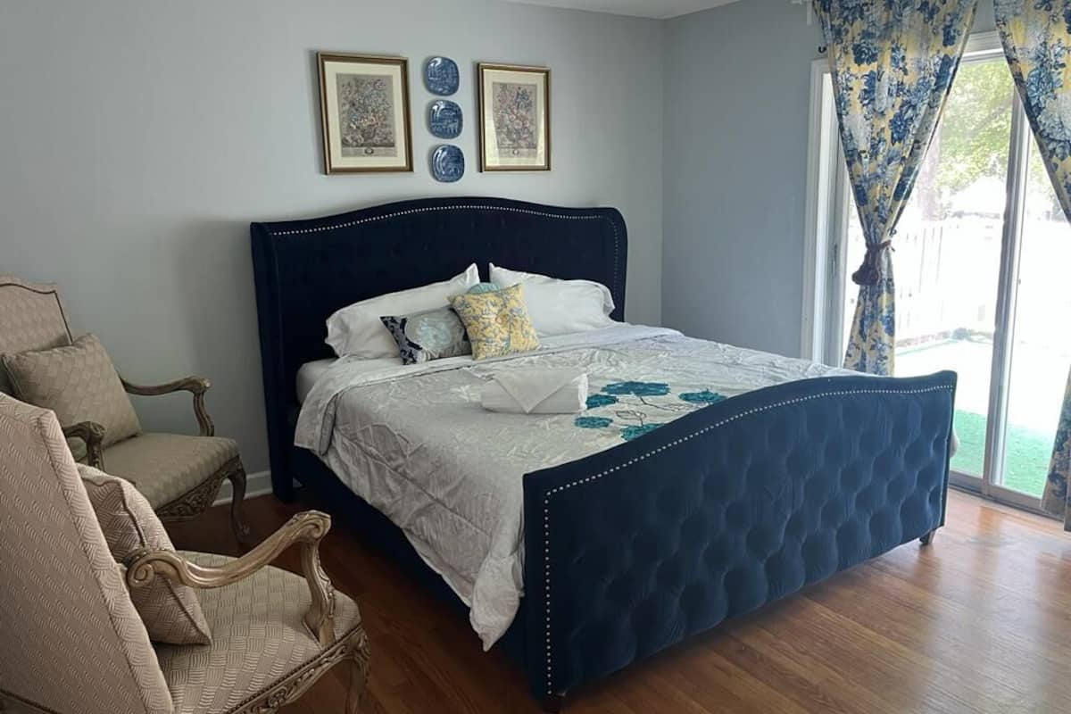 A bedroom with a dark blue upholstered bed, a wooden chair, and a large window with curtains.