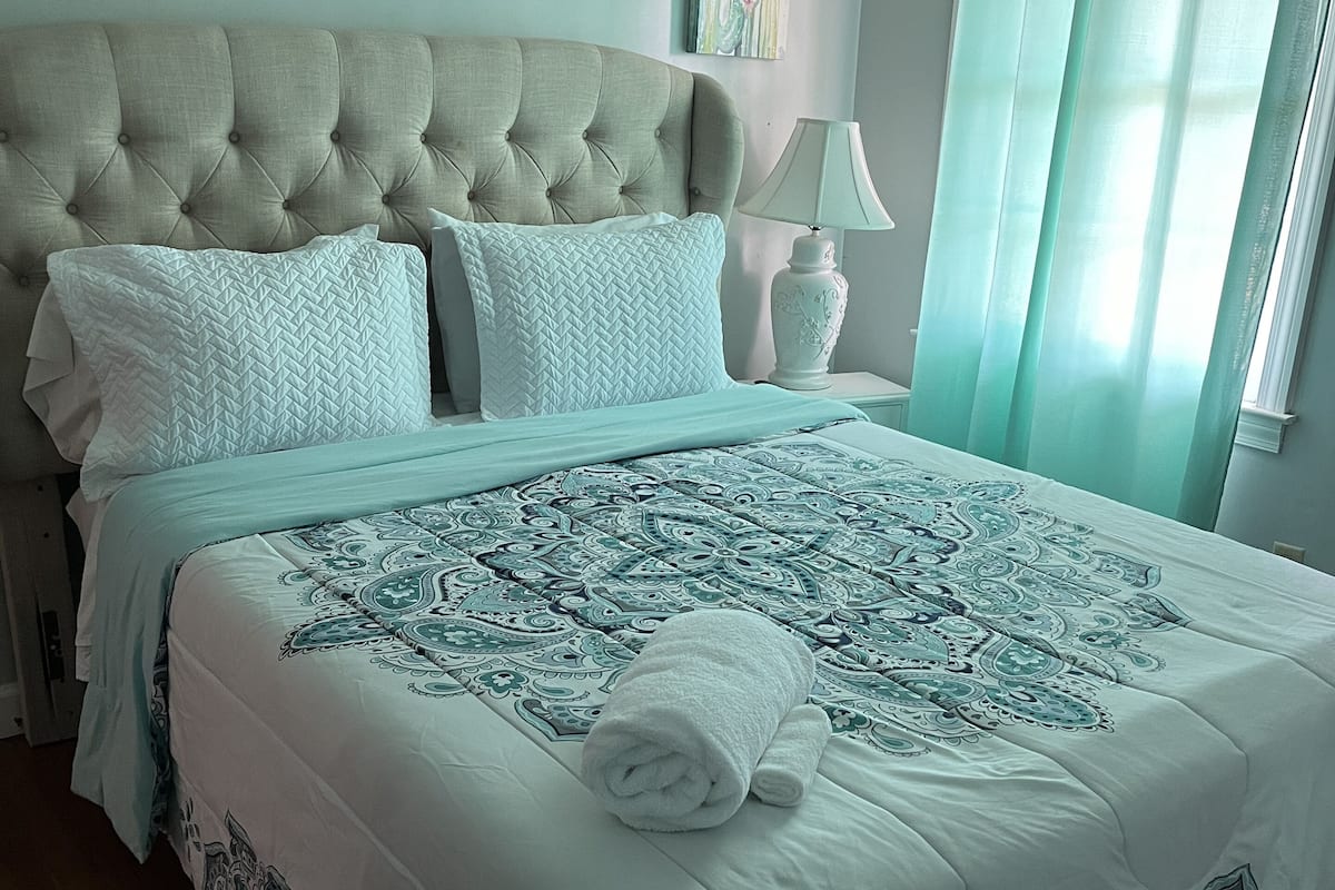 A neatly made bed with a patterned bedspread, a tufted headboard, and a rolled towel on the bed.