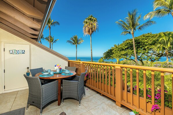 A balcony with wicker chairs and a table set for dining, overlooking a tropical landscape with palm trees and a clear blue sky.