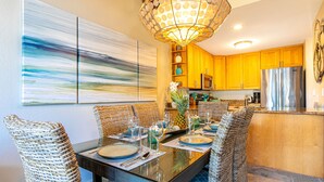 A dining area with a glass table, wicker chairs, and a modern chandelier. - Maui Kamaole #A-210 Panoramic Ocean View, Full A/C, Across Kamaole Bch III (Kihei)