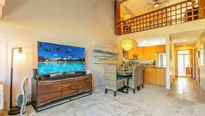 A spacious living area with a large flat-screen TV, a dining table with wicker chairs, a kitchen with wooden cabinets, and a balcony with a wooden railing. - Maui Kamaole #A-210 Panoramic Ocean View, Full A/C, Across Kamaole Bch III (Kihei)