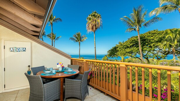 A balcony with wicker chairs and a table set for dining, overlooking a tropical landscape with palm trees and a clear blue sky. - Maui Kamaole #A-210 Panoramic Ocean View, Full A/C, Across Kamaole Bch III (Kihei)