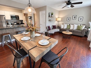 A modern kitchen with stainless steel appliances, a dining area with a wooden table and chairs, a living room with a sofa and a coffee table, and a ceiling fan.