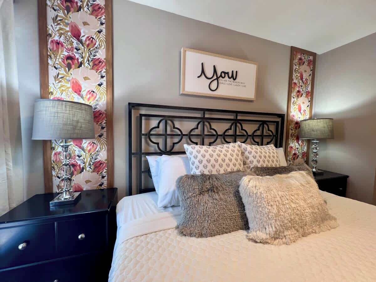 A neatly made bed with a decorative headboard, two bedside tables with lamps, and a floral wallpaper pattern on the walls.