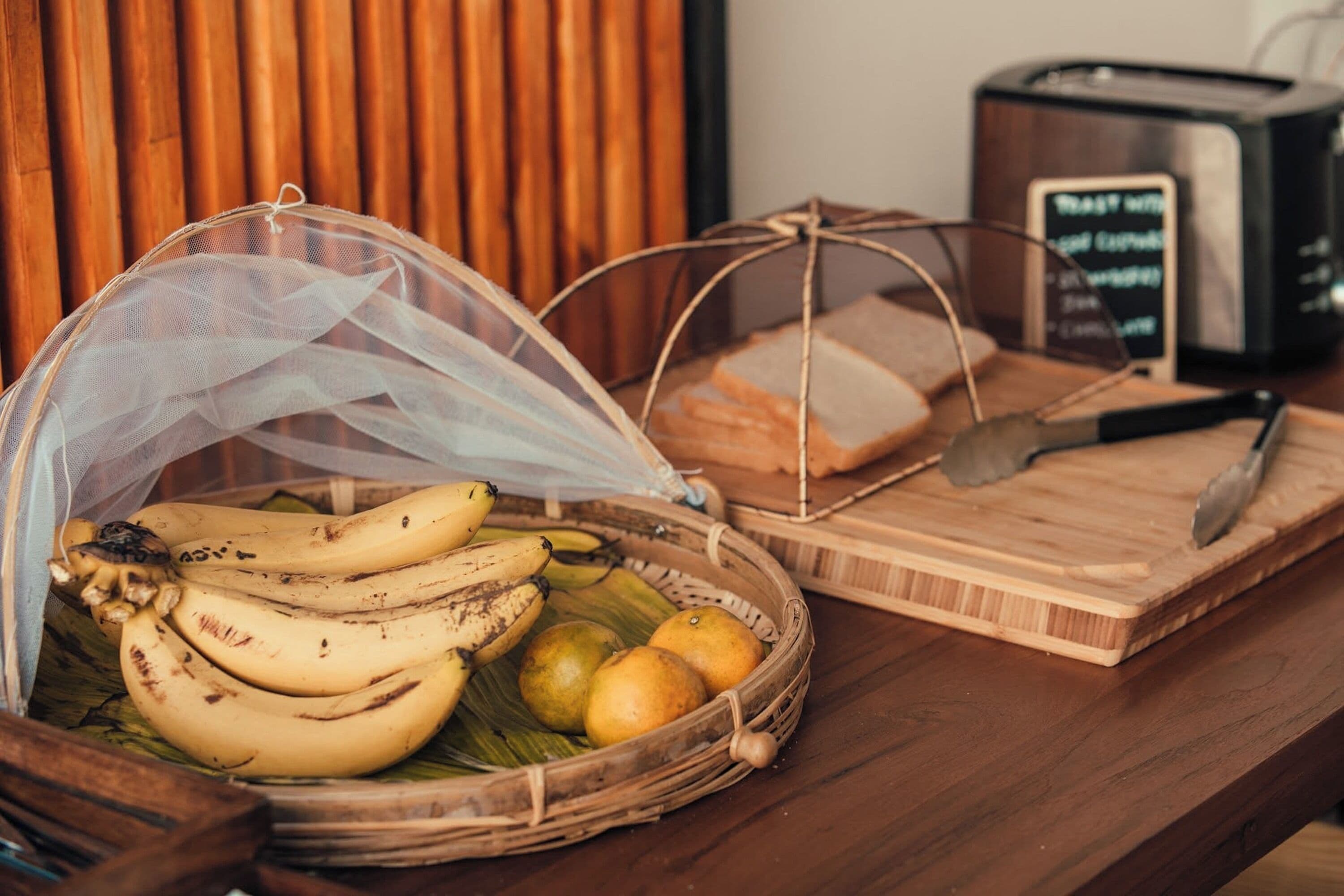 A wooden table with a woven basket containing bananas and oranges, a cutting board with a knife, and a toaster.