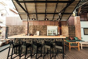 An outdoor bar area with a long counter, black bar stools, a brick wall, and a metal roof structure.