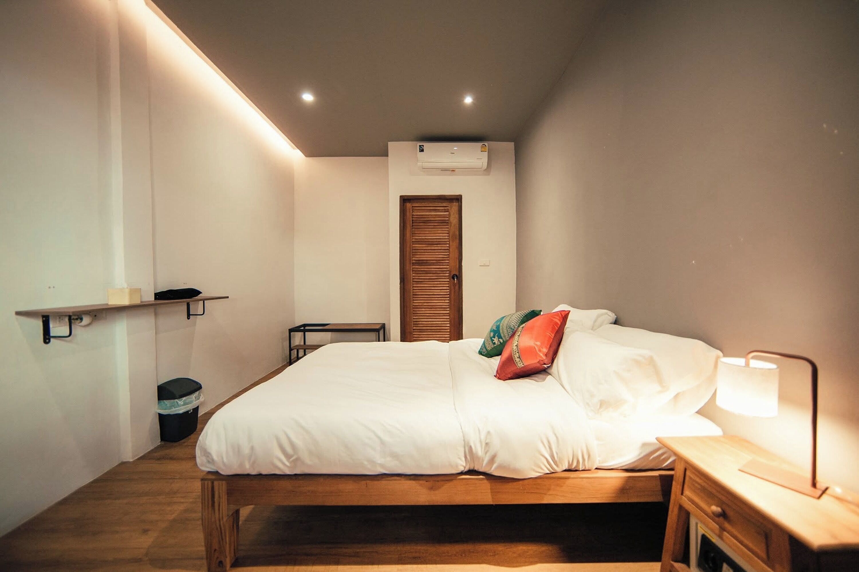 A bedroom with a wooden bed, a bedside table with a lamp, a small table, a trash bin, and a wall-mounted shelf.