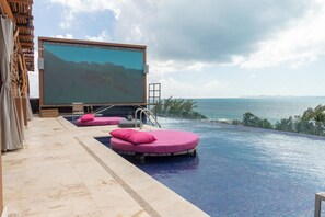 A pool area with a large screen, lounge chairs, and a view of the ocean.