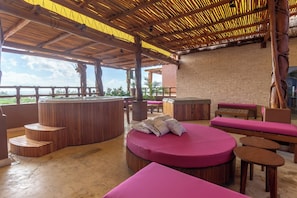 Un espacio lounge al aire libre con techo, bancos de madera y una zona de estar circular con cojines rosados.