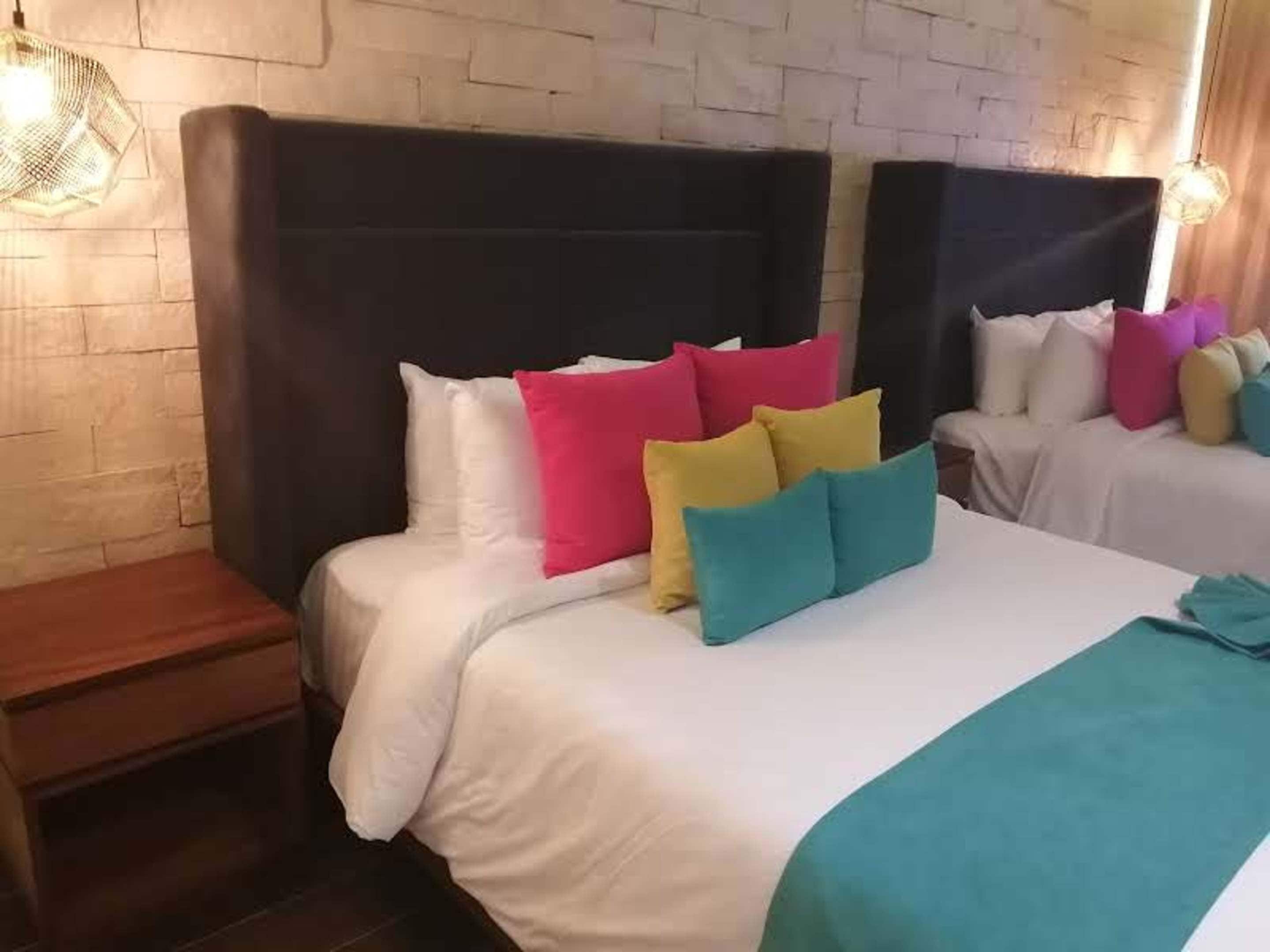 A hotel room with two beds, each with a black headboard, white bedding, and colorful pillows. There is a wooden nightstand and a hanging light fixture.