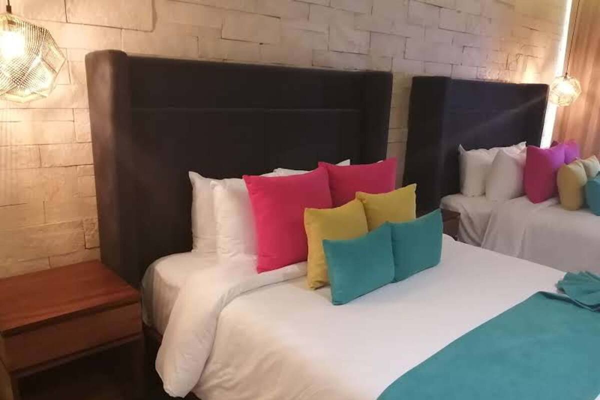 Quarto de hotel com duas camas, cada uma com cabeceira preta, roupa de cama branca e almofadas coloridas. Há uma mesinha de cabeceira de madeira e um candeeiro suspenso.