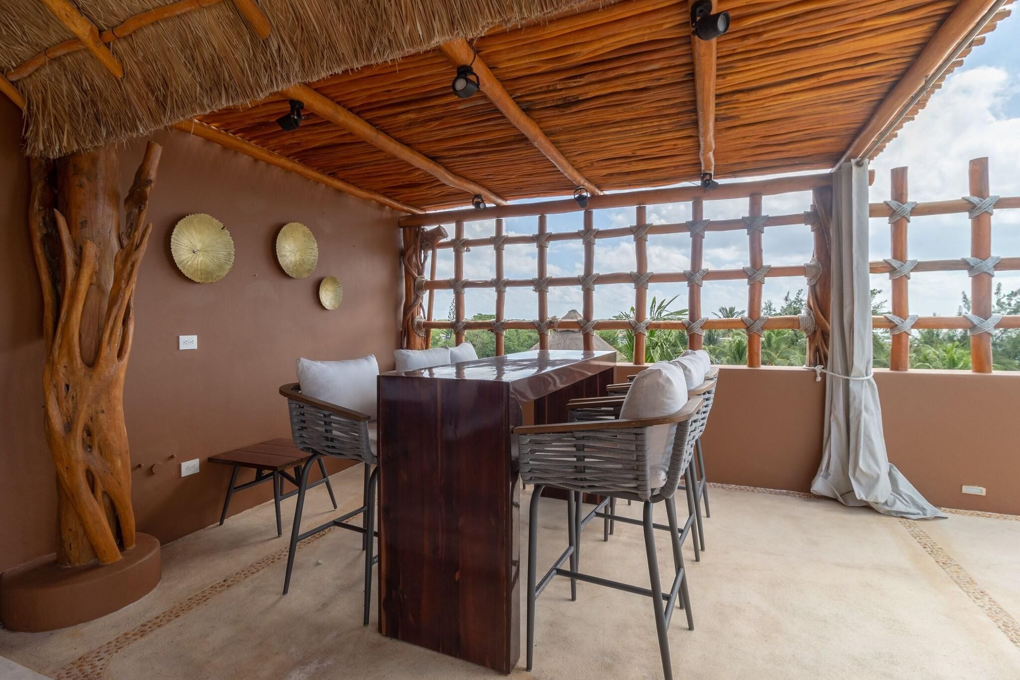 Un espace repas avec une table et des chaises en bois, un toit de chaume et une vue dégagée grâce à de grandes fenêtres.
