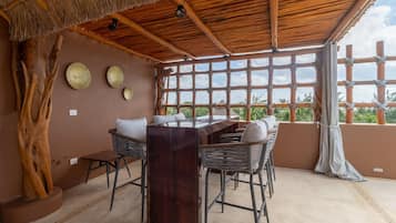 Un espace repas avec une table et des chaises en bois, un toit de chaume et une vue dégagée grâce à de grandes fenêtres.