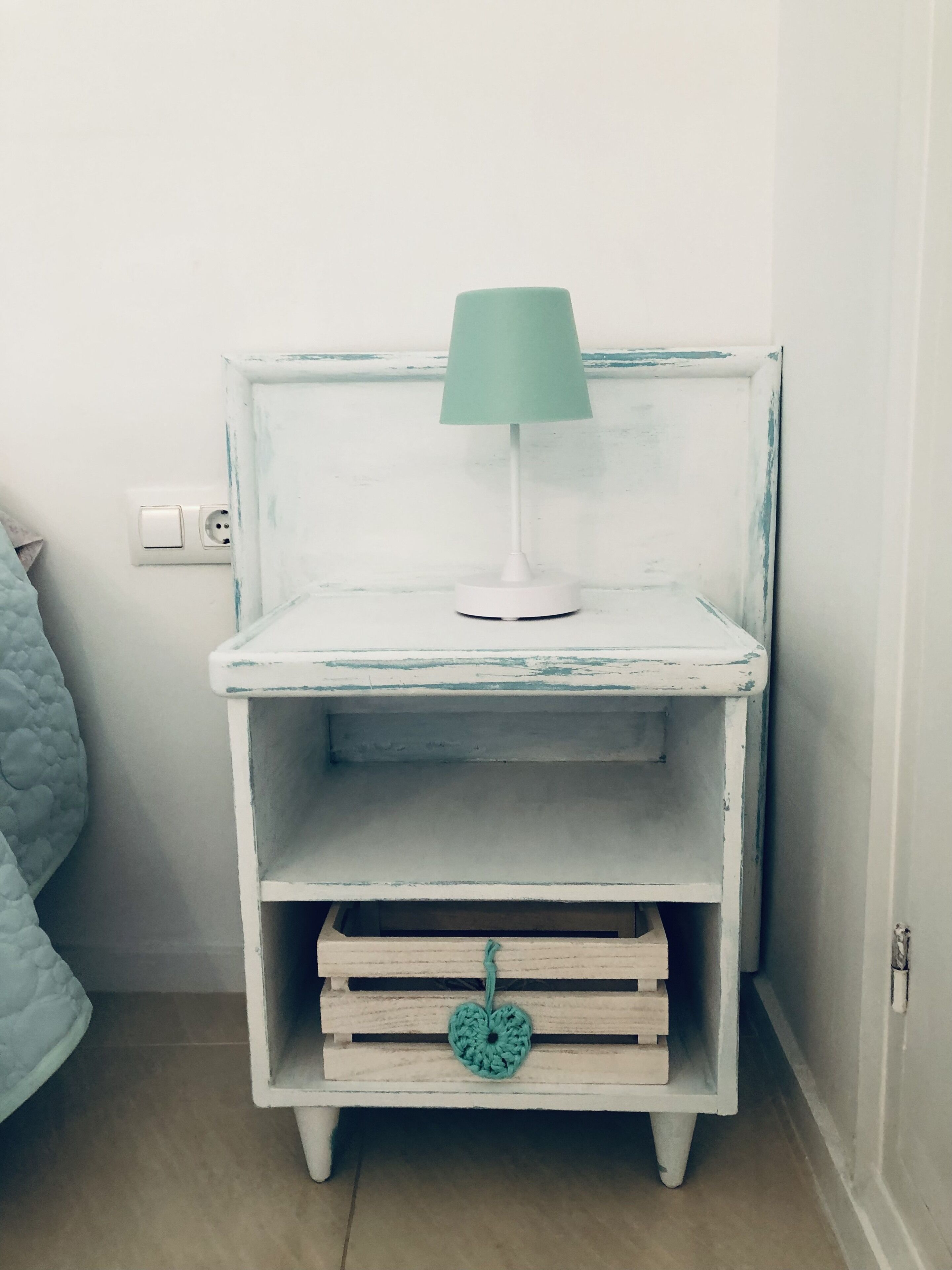 Une table de chevet blanche, surmontée d’une lampe turquoise, avec un tiroir orné d’un cœur turquoise et une prise électrique au mur.
