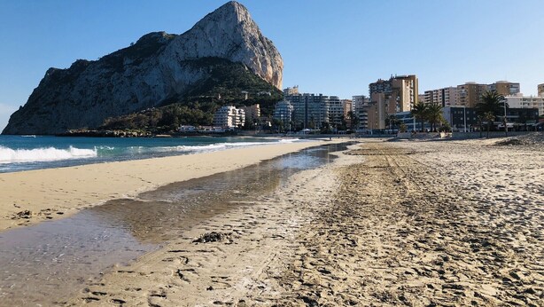 En strand med en stor klippformation, en sandstrand och byggnader i bakgrunden.