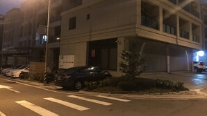 Un edificio de varios pisos con fachada plana, una entrada protegida por una reja y un estacionamiento con varios automóviles.
