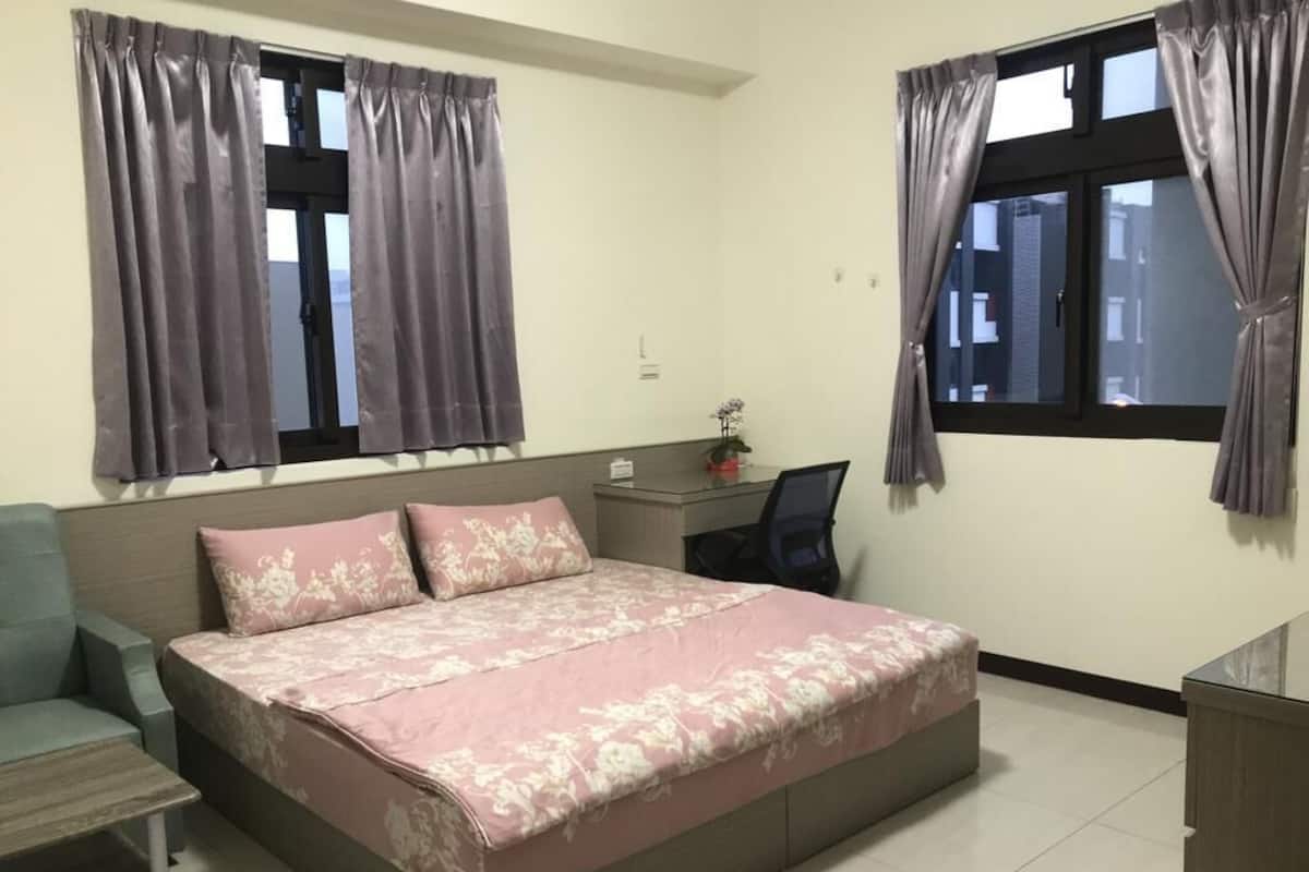 Un dormitorio con cama, escritorio, silla y dos ventanas con cortinas.