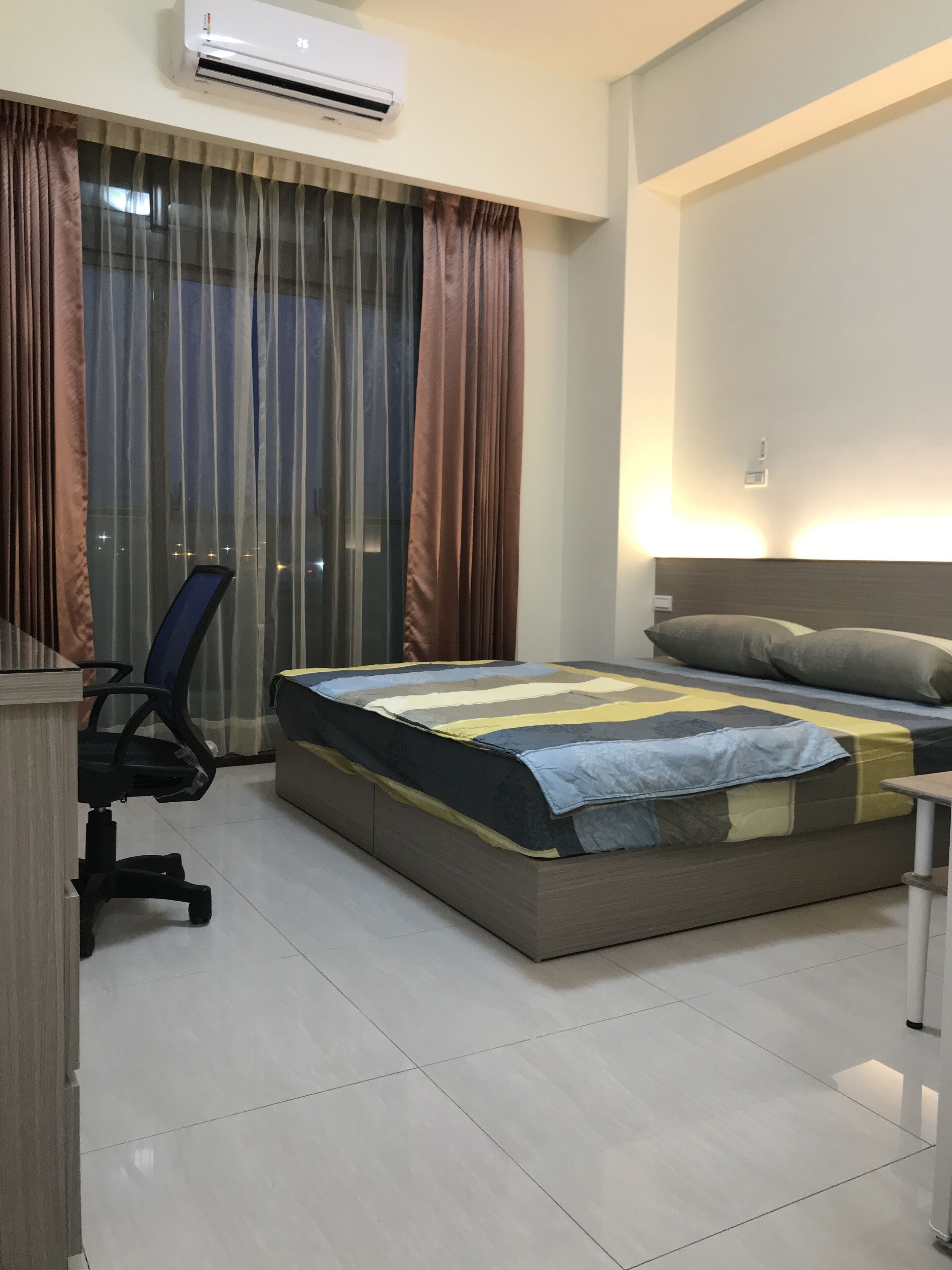 A hotel room with a bed, a desk, an office chair, a window with curtains, and an air conditioning unit.