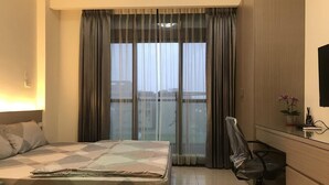 Habitación de hotel con cama, escritorio, silla, planta en maceta y vistas a la ciudad a través de amplias ventanas.
