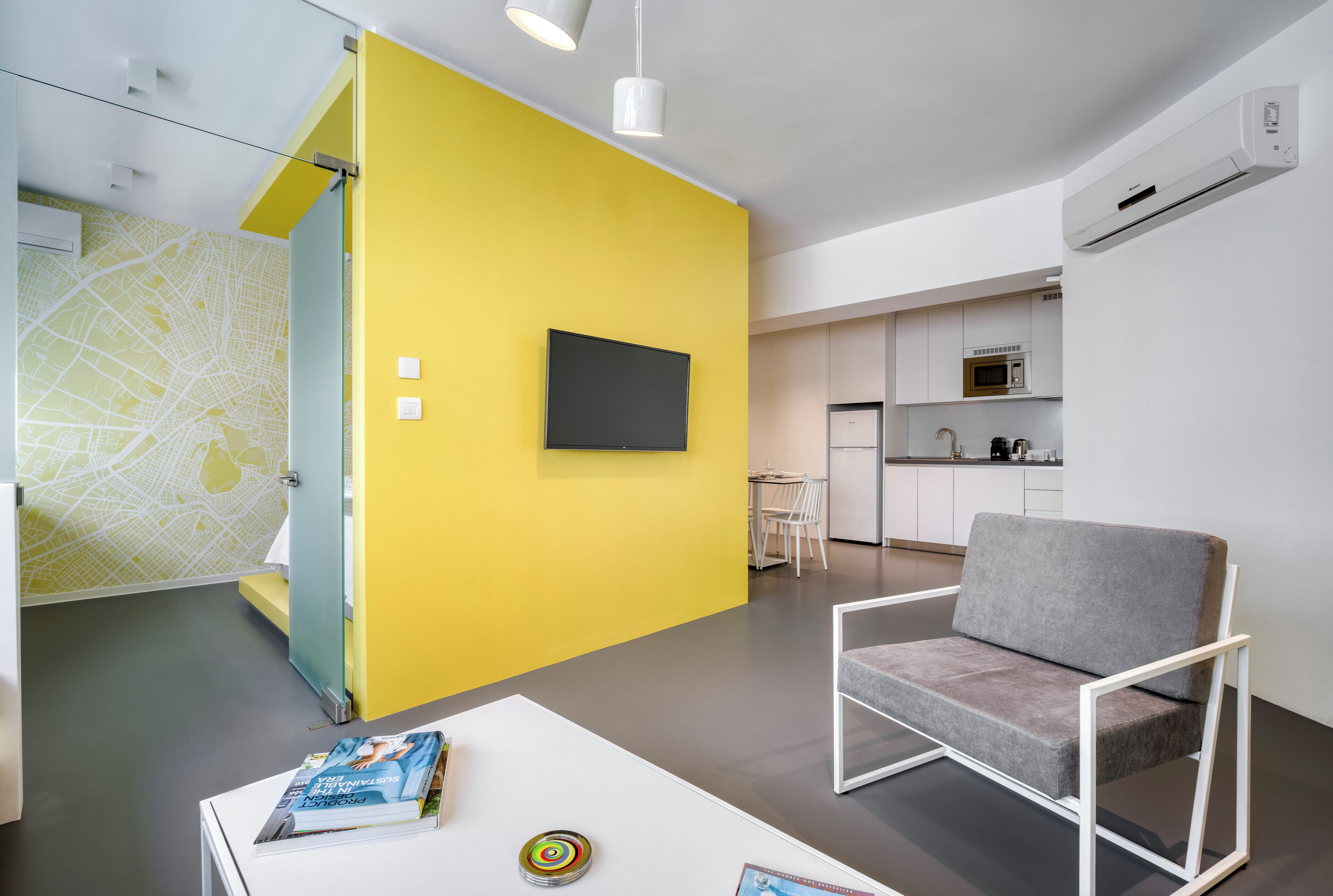 A modern living room with a yellow accent wall, a grey sofa, a flat-screen TV, and a white coffee table with books and a decorative item.