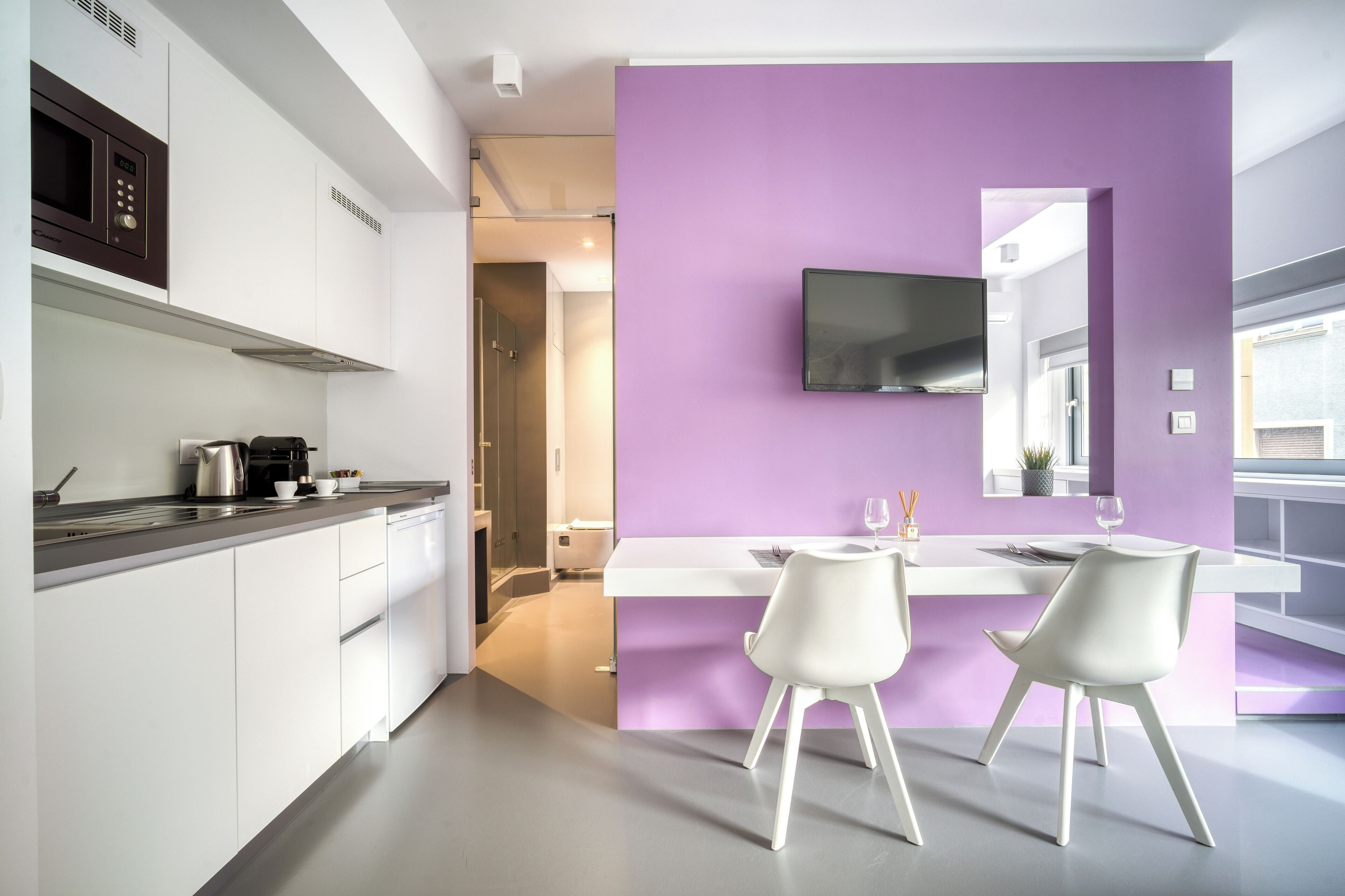 A modern kitchen with white cabinets, a purple accent wall, and a flat-screen TV mounted above the counter.