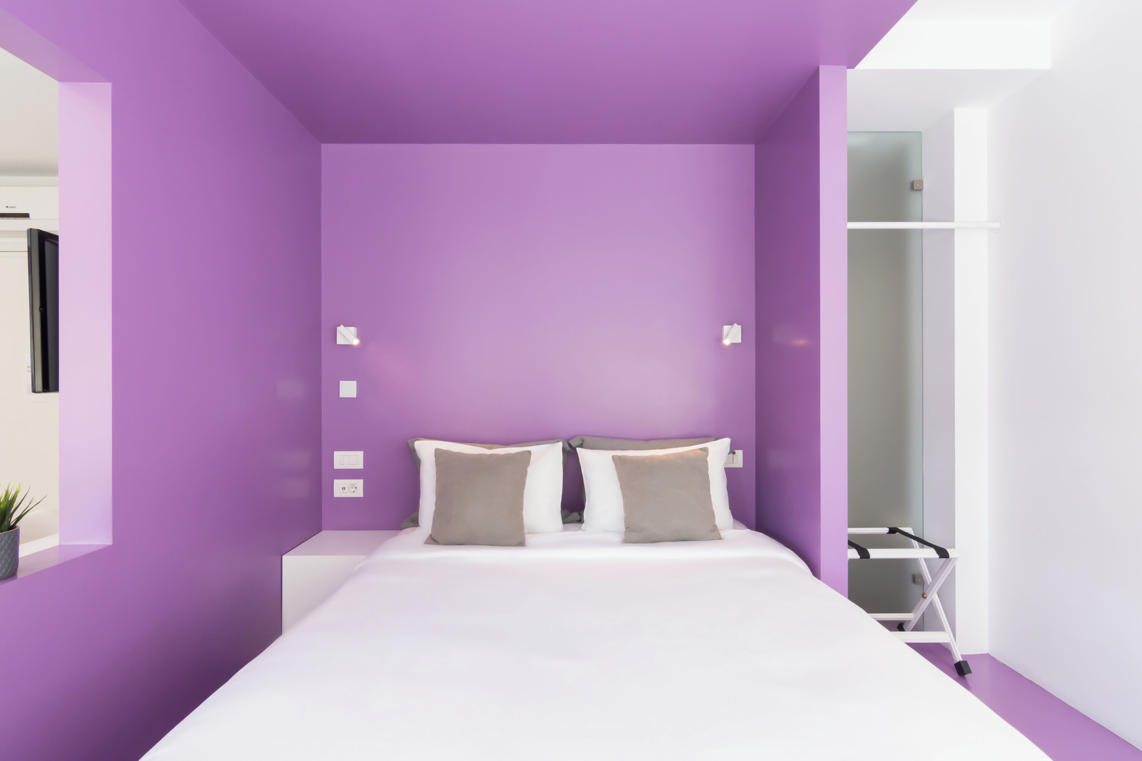 Een moderne slaapkamer met een paarse muur, een wit bed en een klein nachtkastje.