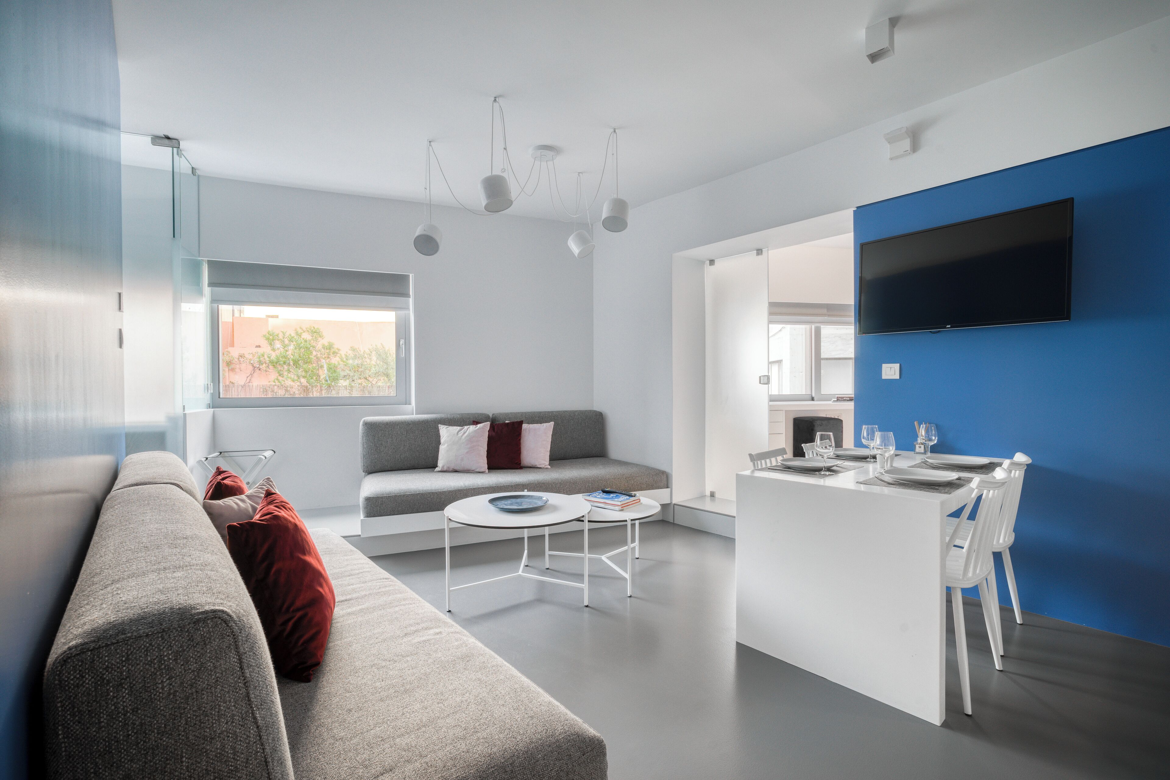 Una sala de estar moderna con un sofá gris, una mesita redonda y una cocina con una pared azul de acento y un televisor de pantalla plana.
