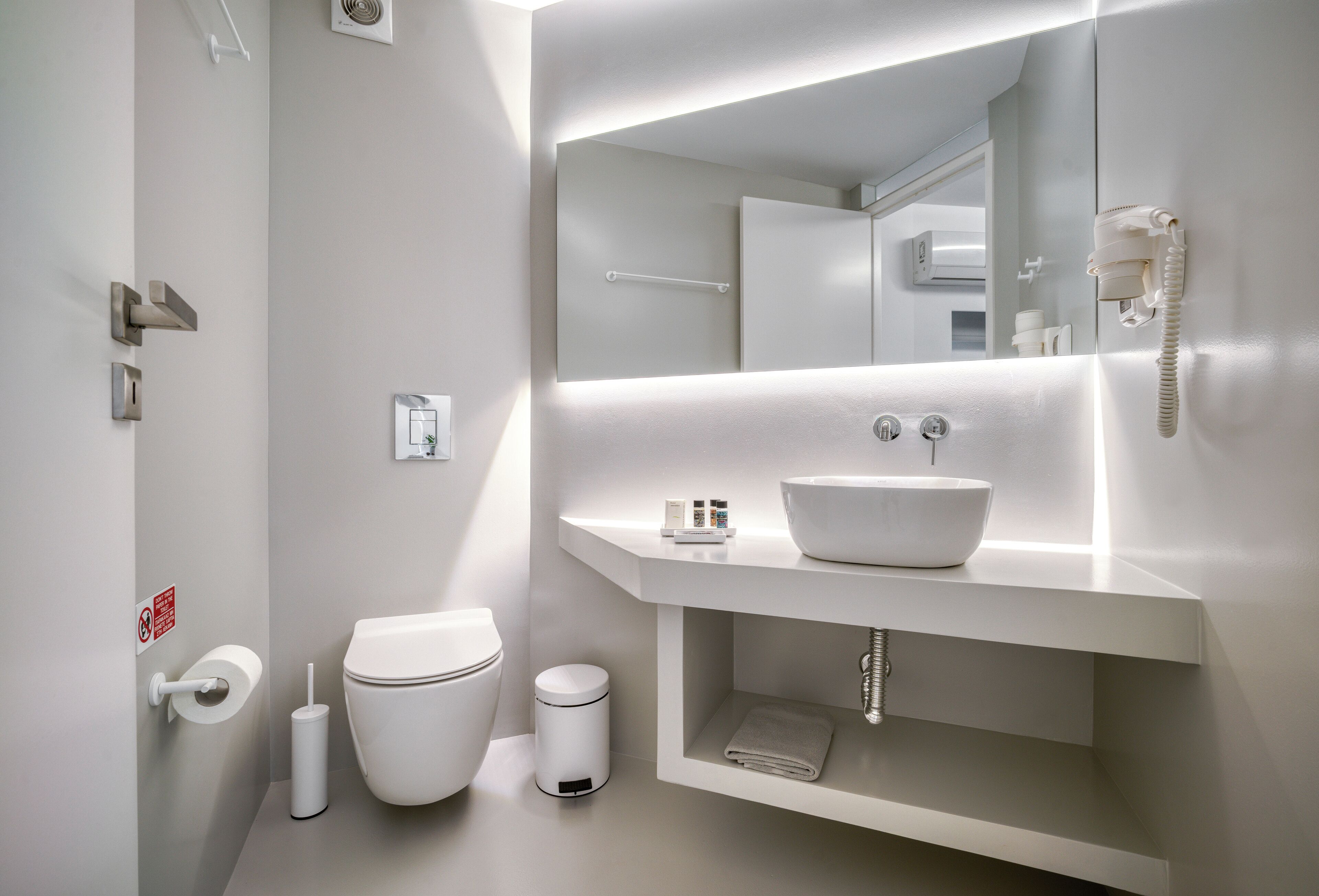 A modern bathroom with a white sink, toilet, and bidet. There is a mirror above the sink and a telephone mounted on the wall.