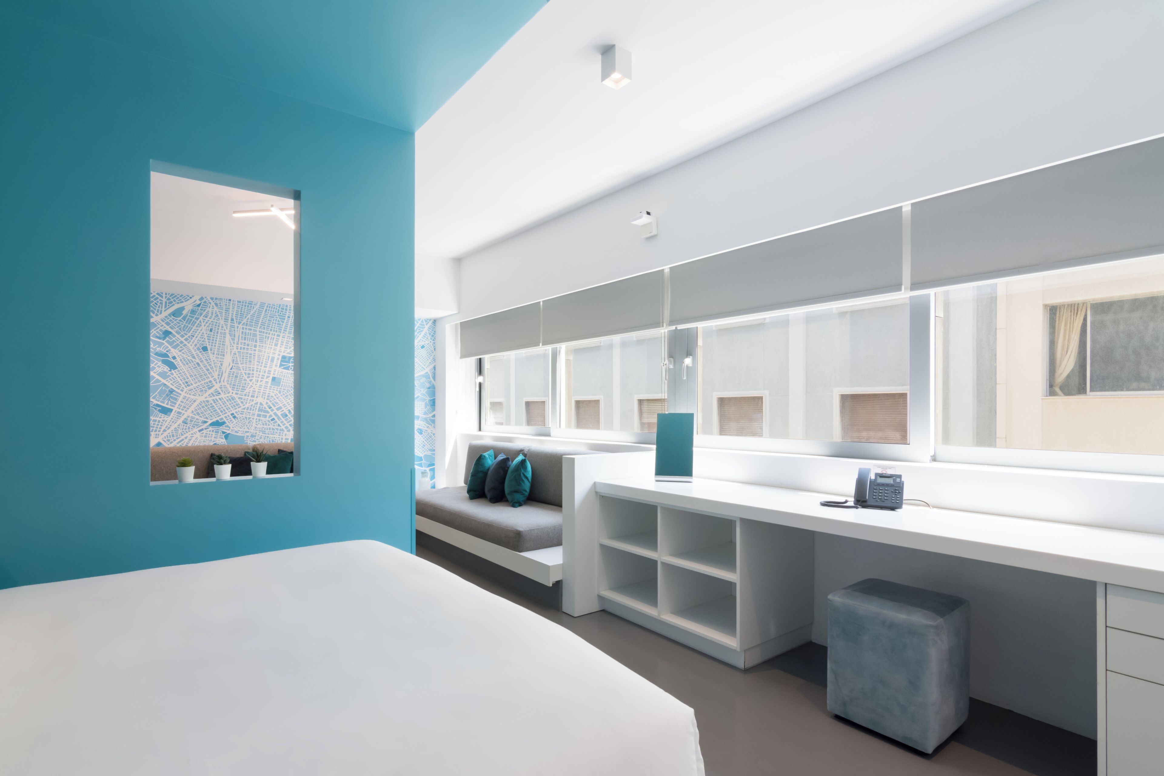 A modern hotel room with a large bed, a desk with storage, a seating area, and a large window.