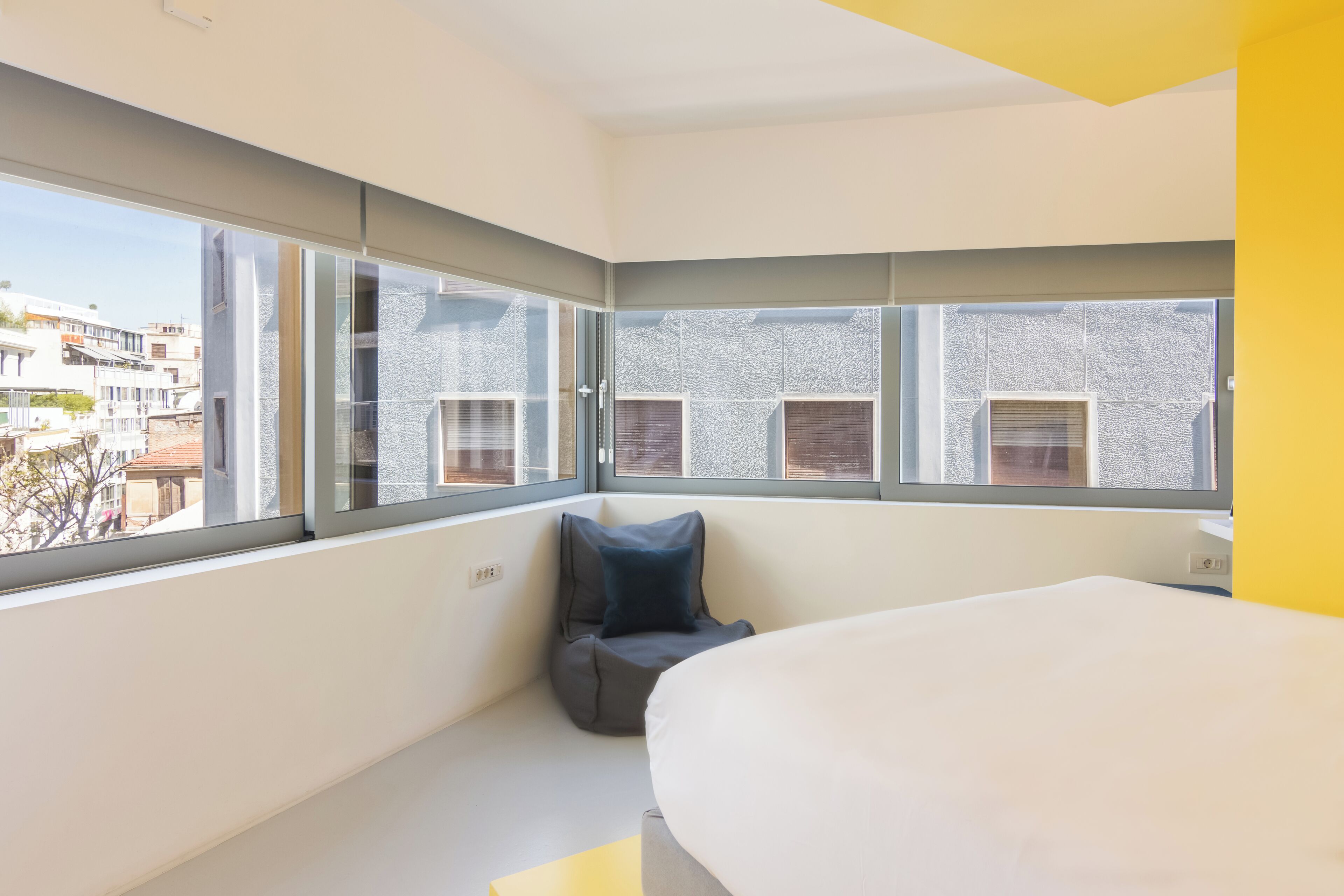 A modern hotel room with a large bed, a seating area with a blue cushion, and a view of the city through large windows.