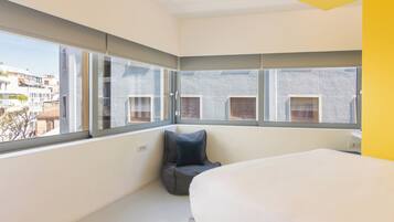 A modern hotel room with a large bed, a seating area with a blue cushion, and a view of the city through large windows.