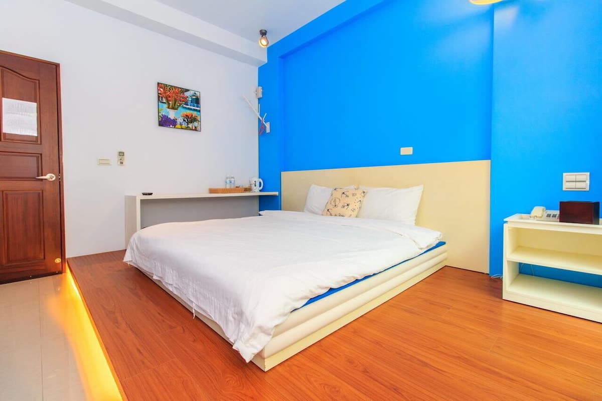 A bedroom with a bed, a wooden floor, a blue accent wall, a small bedside table, and a wall-mounted light fixture.