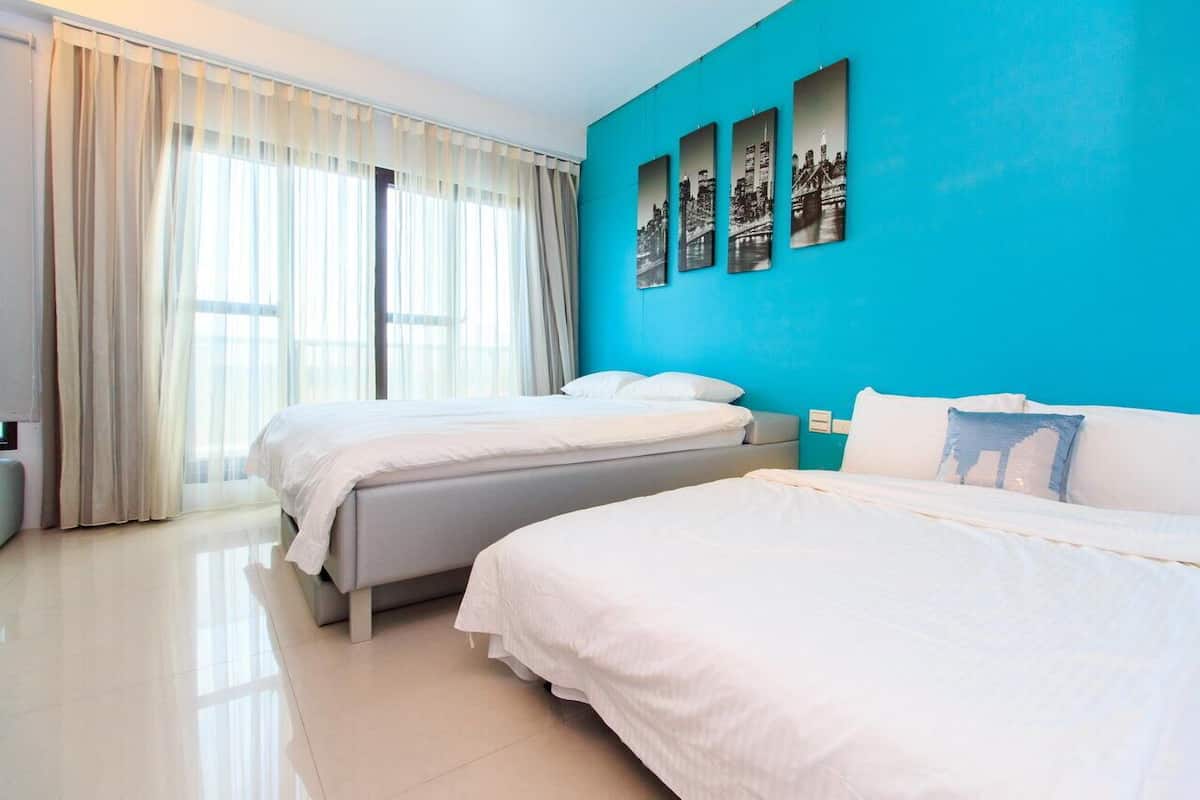 A hotel room with two beds, a large window with sheer curtains, and a bright blue accent wall with black-and-white cityscape pictures.