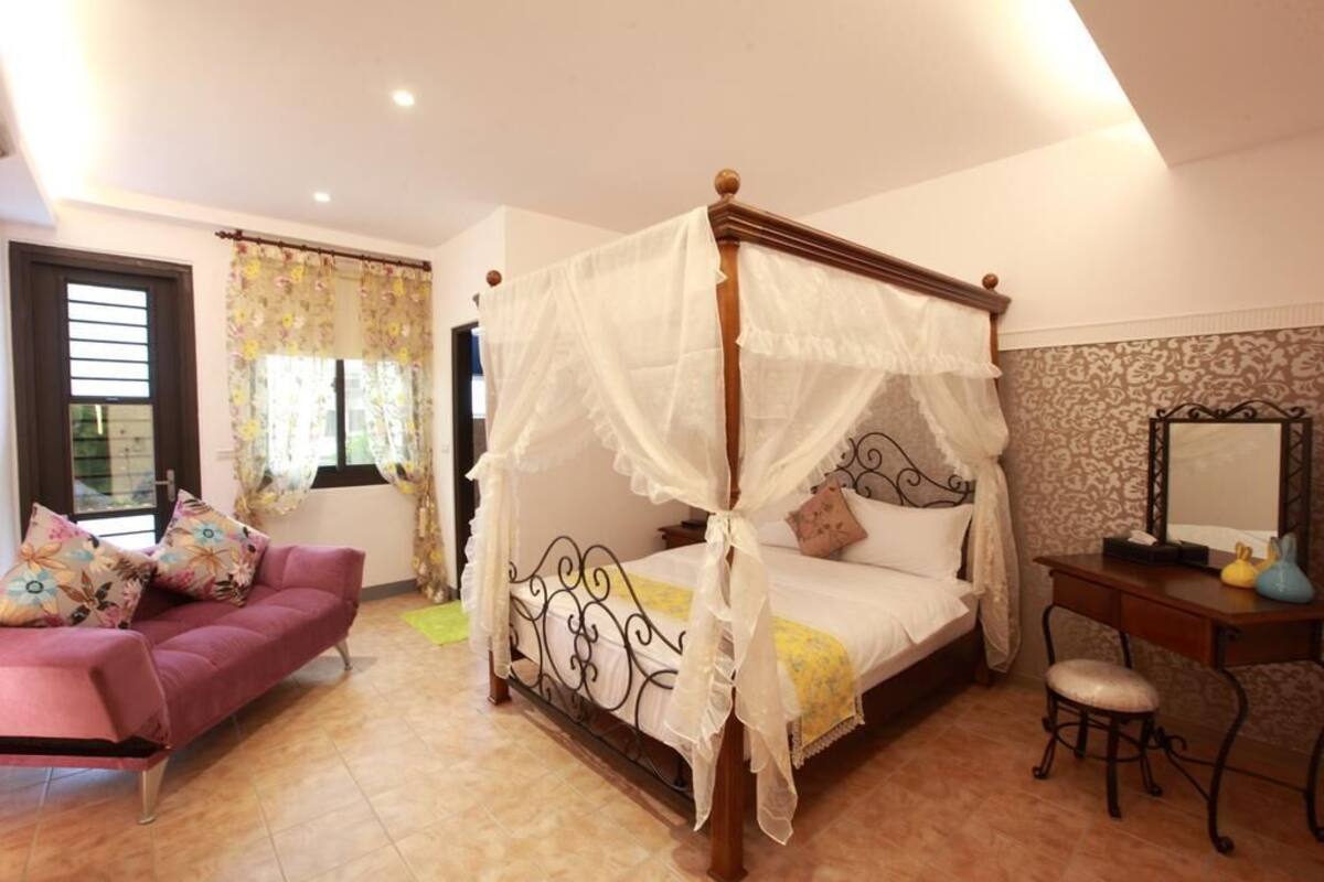 A four-poster bed with a canopy, a sofa with floral cushions, a desk with a mirror, and a window with curtains.