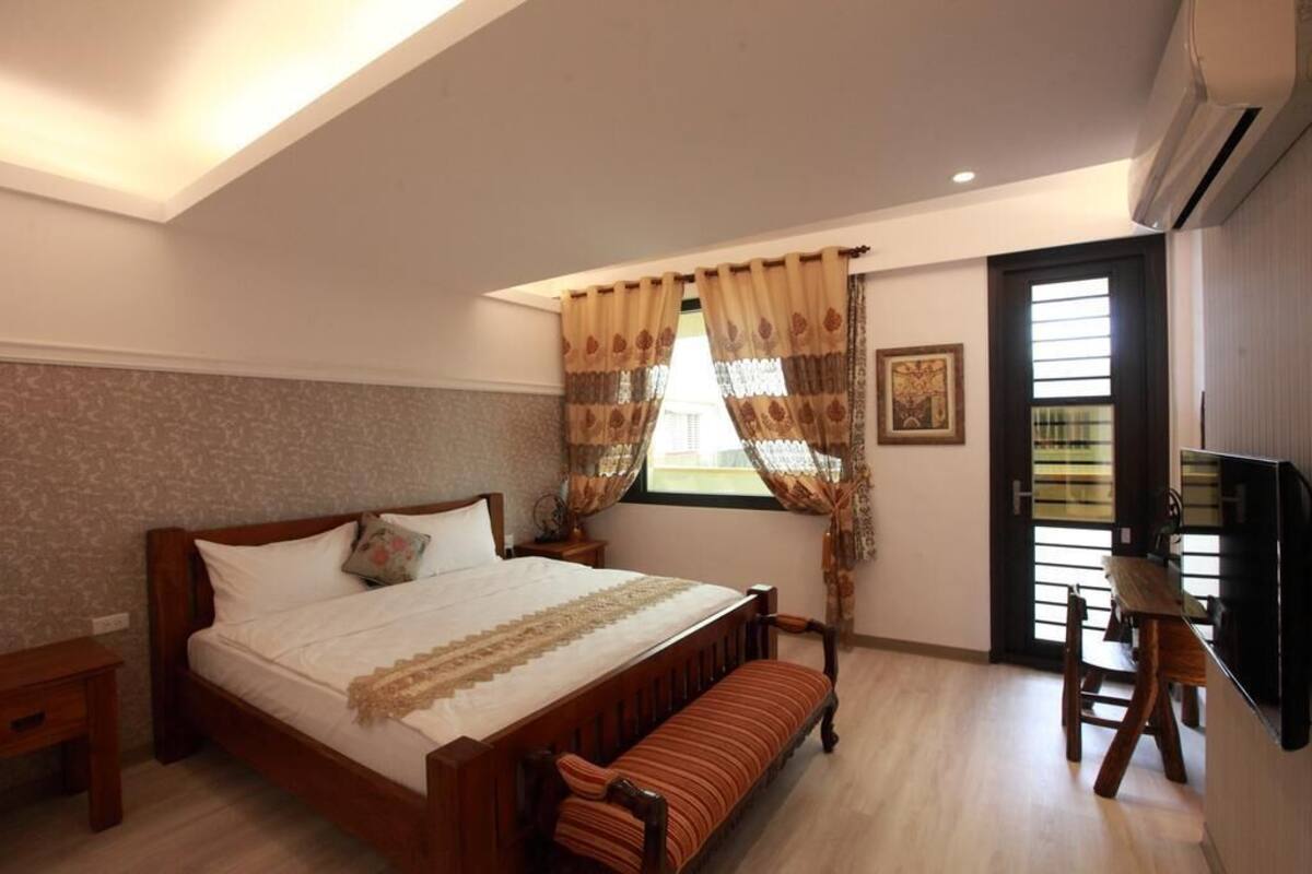 A bedroom with a bed, bench, bedside table, curtains, a framed picture, and a TV.