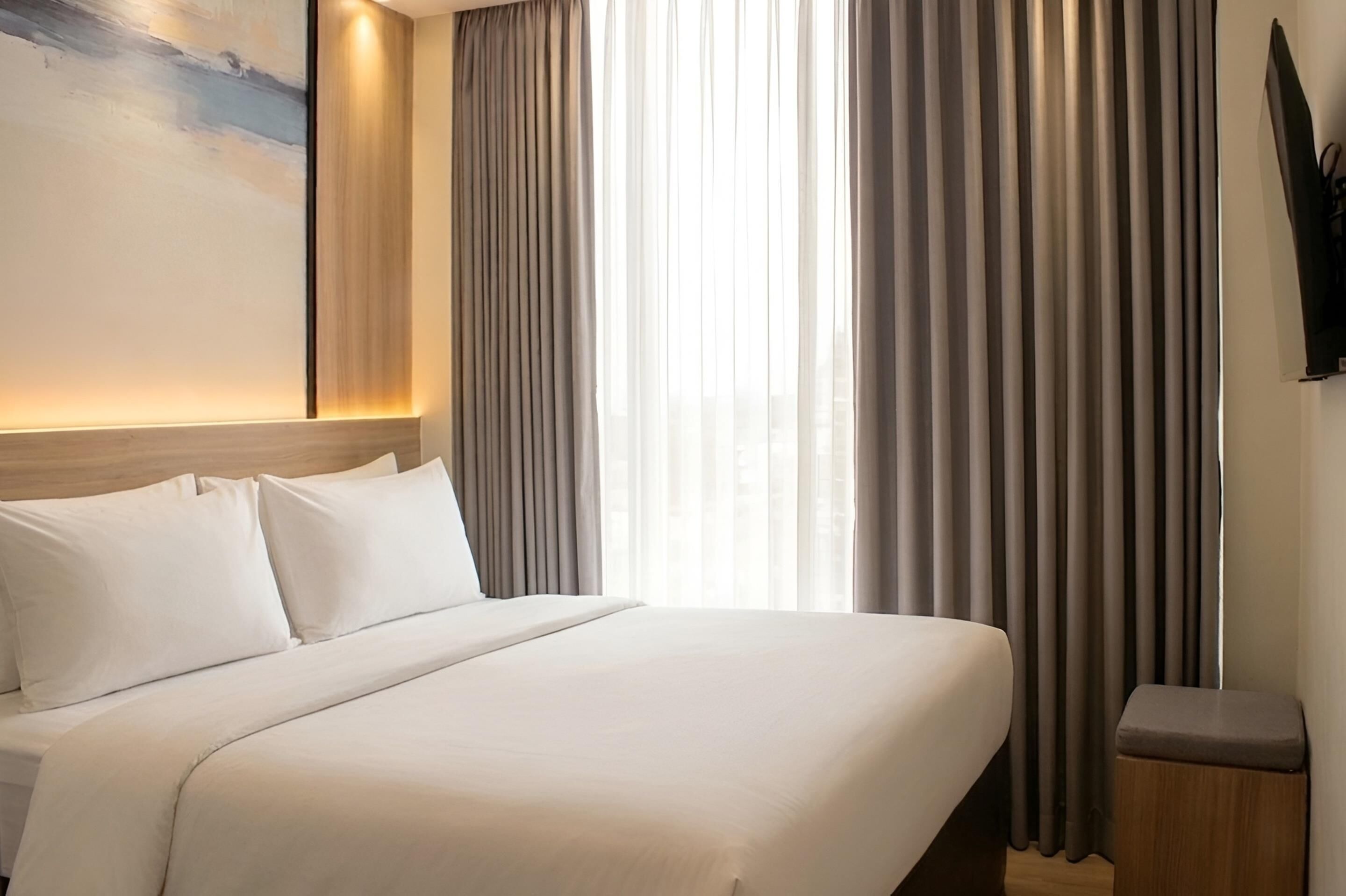 A hotel room with a bed, curtains, a wall-mounted TV, and a painting.