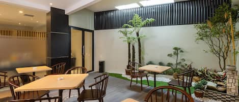 Un espace de restauration intérieur moderne, avec des tables et des chaises en bois, un petit jardin agrémenté de plantes et un mur en briques, ainsi qu’une lucarne au-dessus.