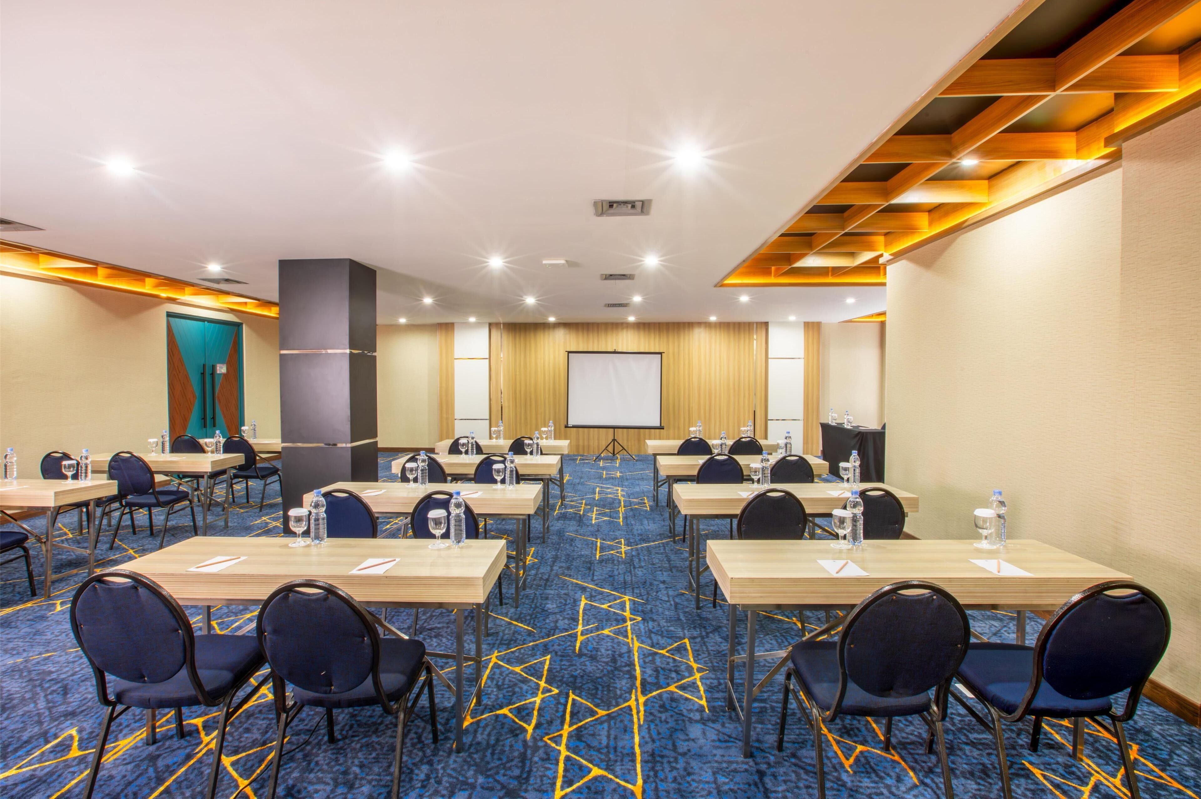 A conference room with multiple tables and chairs, a projector screen, and a carpeted floor.