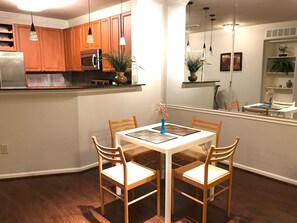 A modern kitchen with wooden cabinets, a stainless steel refrigerator, and a microwave. There is a dining table with four chairs and a vase of flowers.