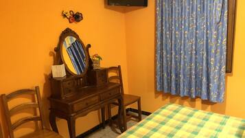 Une pièce comprenant une commode en bois, un miroir, un lit recouvert d’une courtepointe à carreaux, un téléviseur fixé au mur et un rideau à motifs floraux.