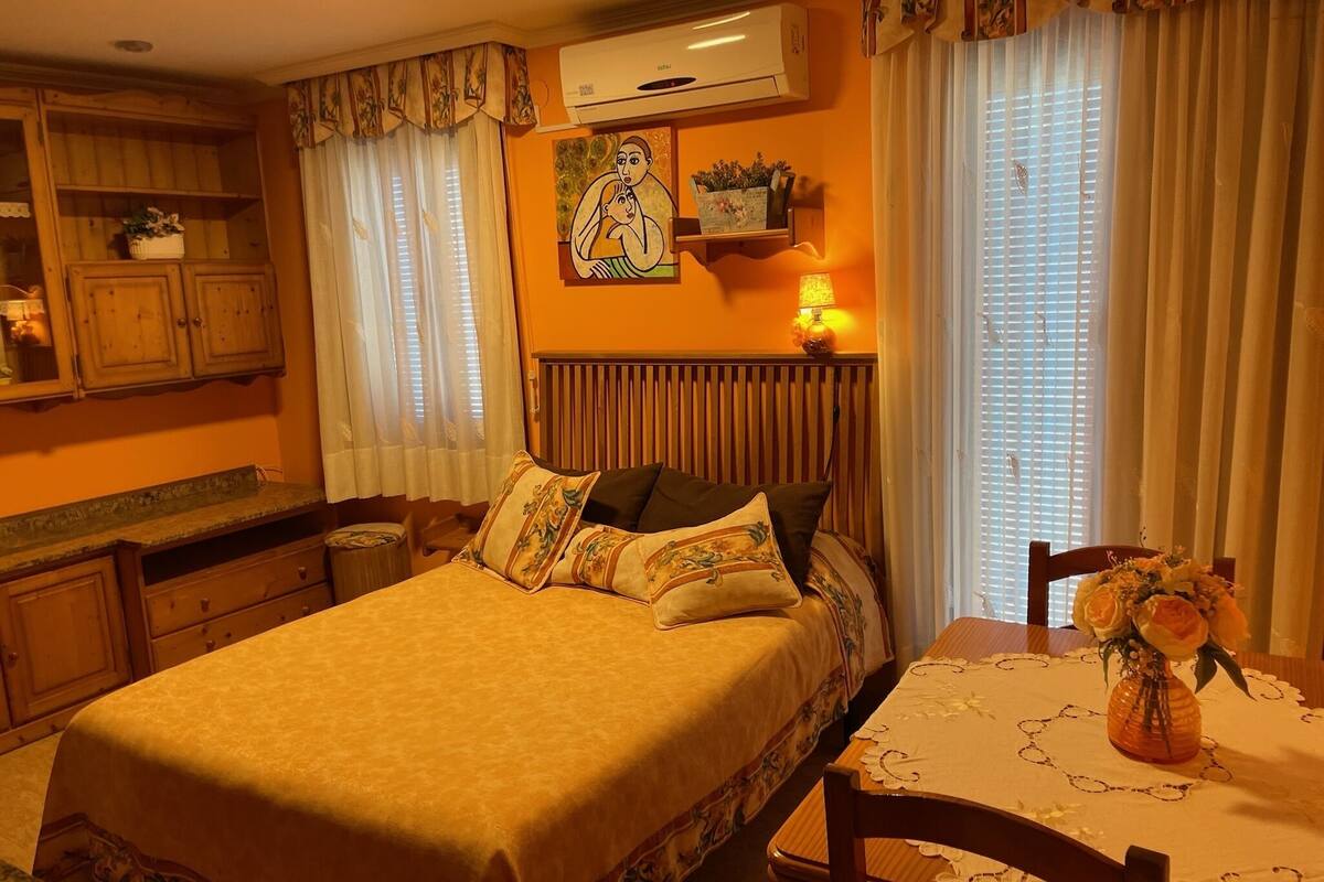 A cozy bedroom with a bed, a bedside table with a vase of flowers, a wall-mounted air conditioner, and a window with curtains.