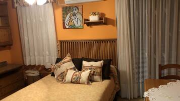 A bedroom with a bed, pillows, a nightstand, a dresser, a ceiling fan, an air conditioner, a painting, and curtains.
