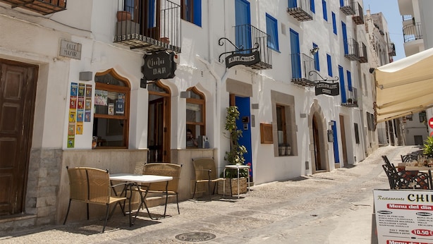 A white building with blue shutters and multiple balconies. There are outdoor tables and chairs in front.