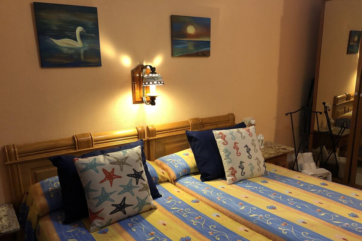 A hotel room with a bed featuring striped bedding, star and seahorse patterned pillows, a nightlight, and two paintings on the wall.