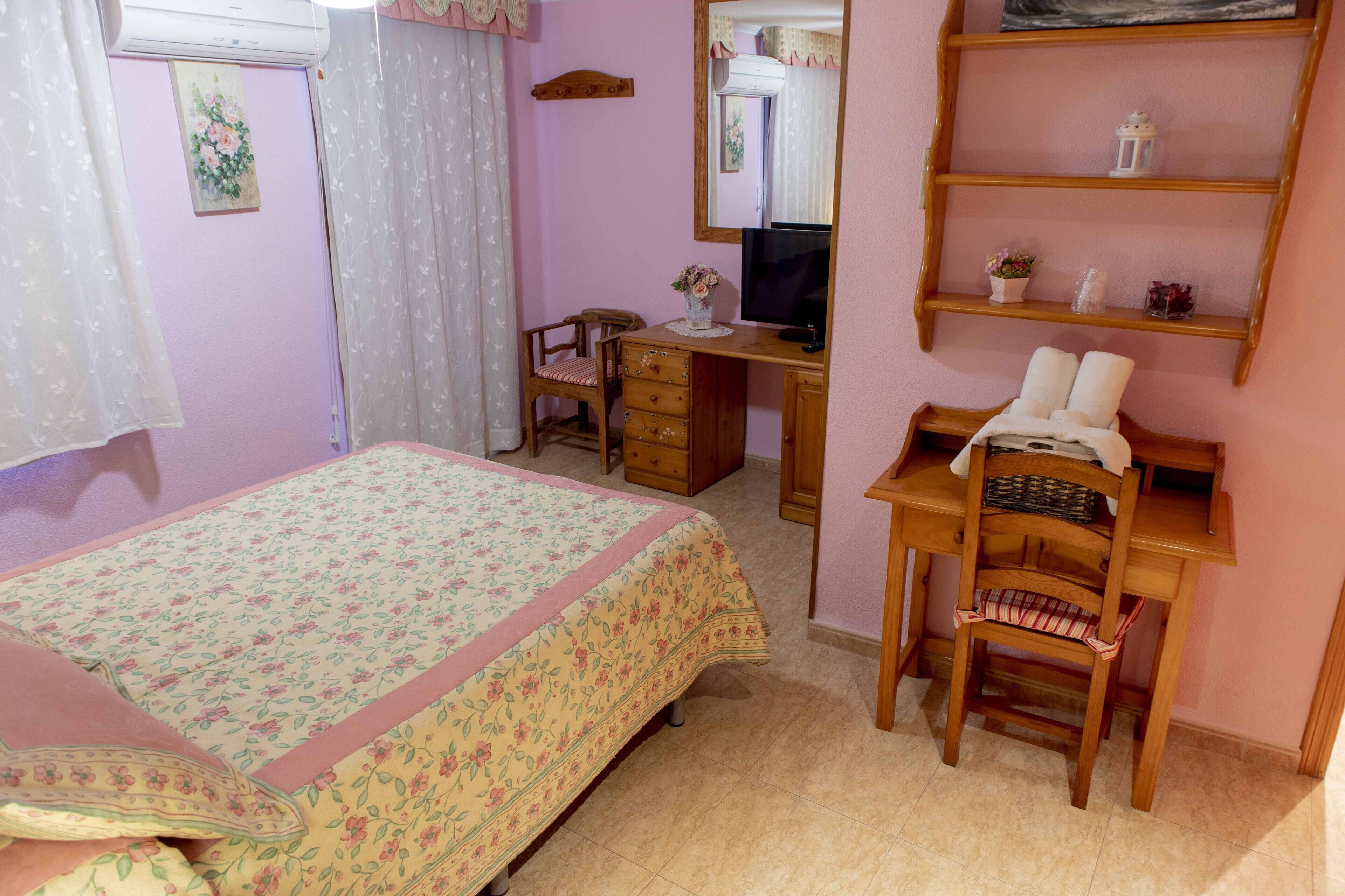 Een slaapkamer met een bed, een bureau, een stoel, een televisie en een plank met decoratieve objecten.