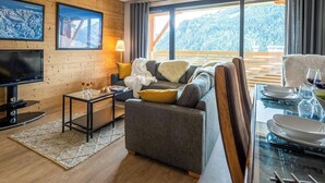 Una sala de estar acogedora con un sofá, un televisor, una mesa de vidrio dispuesta para comer y vistas a una montaña boscosa.